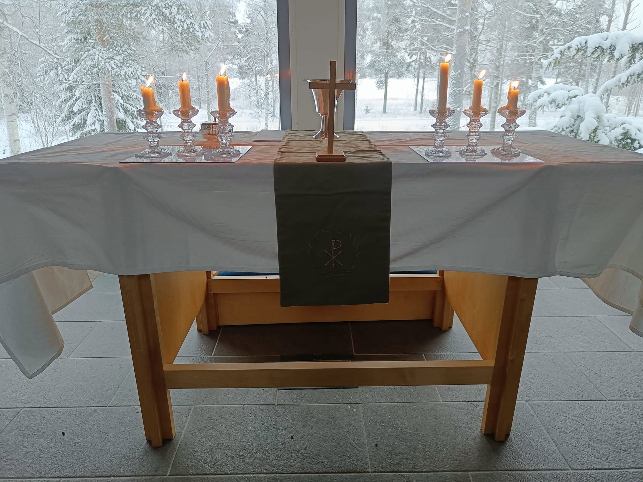 Alttari jossa kynttilöitä ja risti. Taustalla talvimaisema ikunan läpi.