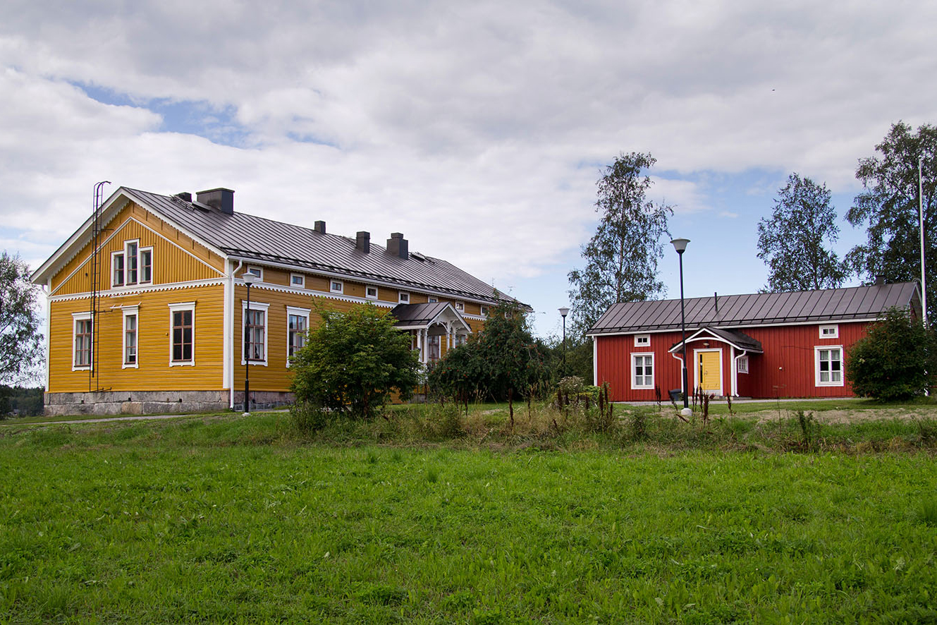 Pihapiiri jossa keltainen puuraknteinen pappilla, jossa tummanruskea peltikatto. Samalla pihalla on pienempi punainen puurakennus.